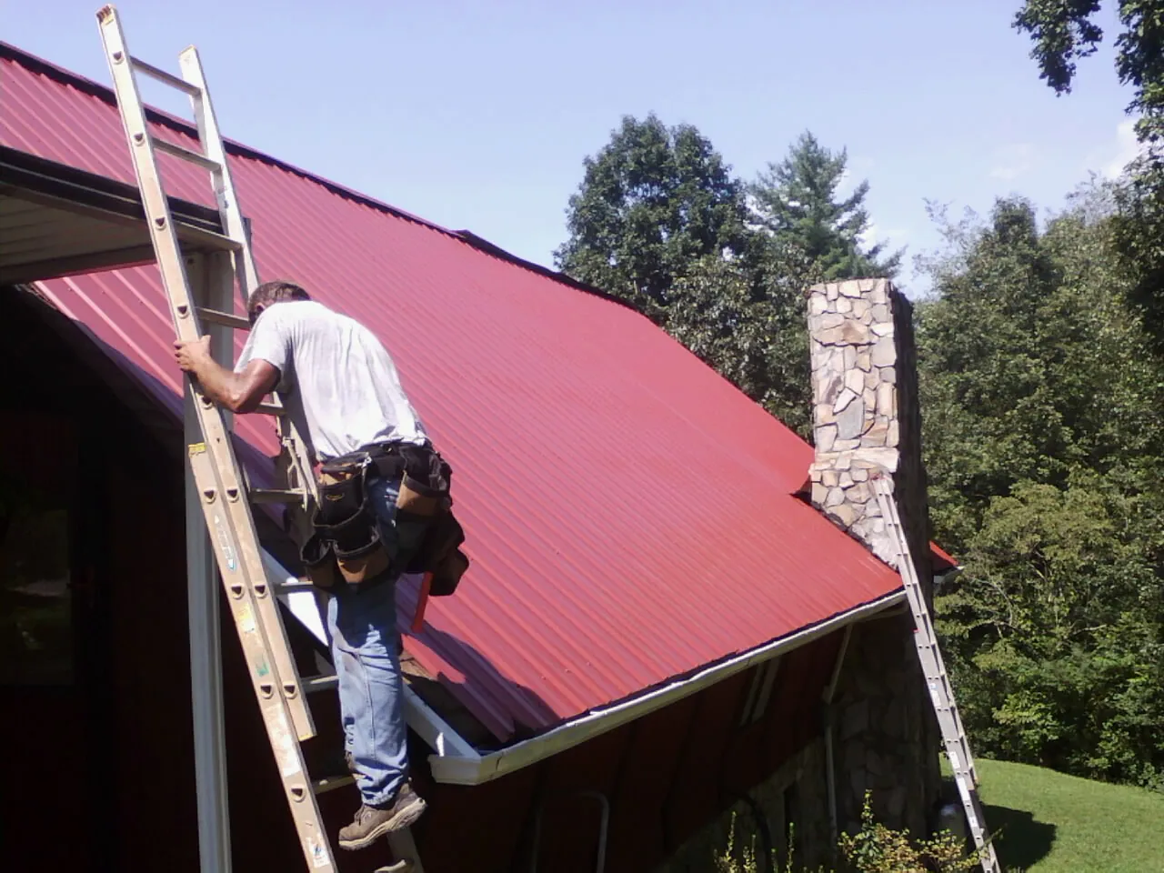 Roofer on a ladder beside a red metal roof and stone chimney — Soffit & Fascia Repair by Roofing Brothers in Sandwich